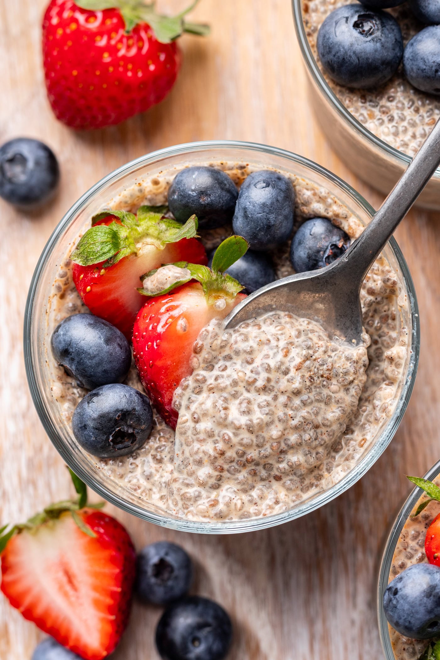A glass bowl of chia pudding topped with fresh strawberries and blueberries. A spoon is scooping out some pudding, and more berries are scattered on the wooden surface nearby.