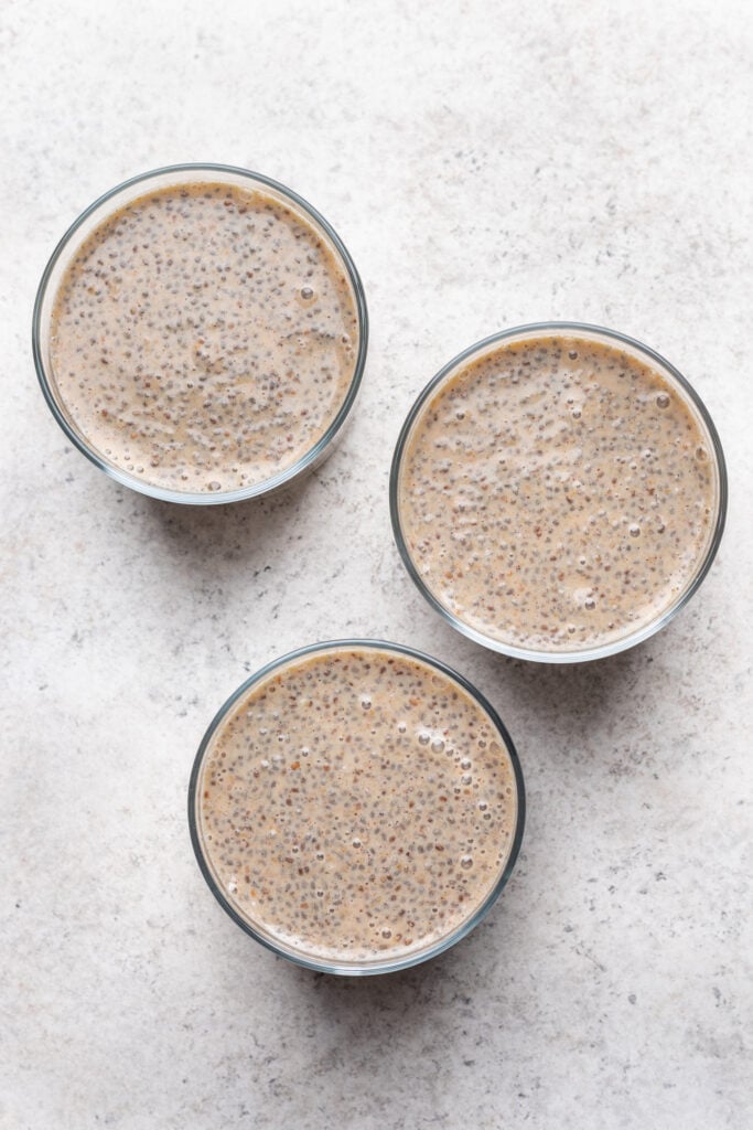 Three glass bowls filled with chia seed pudding arranged in a triangular pattern on a light, textured surface. The pudding has a creamy beige color with visible chia seeds throughout.