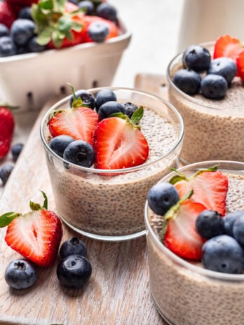 Three glass cups filled with chia pudding topped with fresh strawberries and blueberries, with extra berries scattered on a wooden surface and a basket of strawberries in the background.