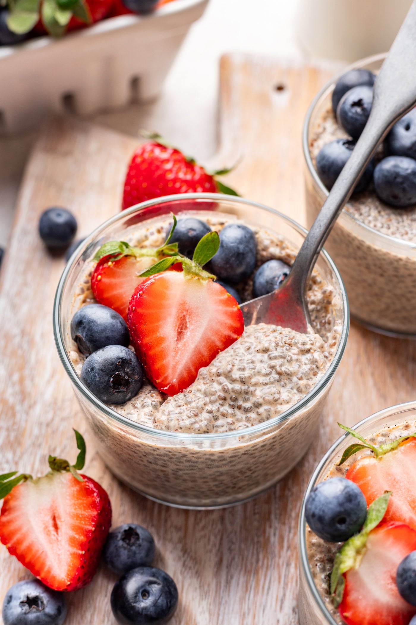 A glass dish of chia pudding topped with fresh blueberries and sliced strawberries sits on a wooden board, with a spoon inside. More berries are scattered around the dish.