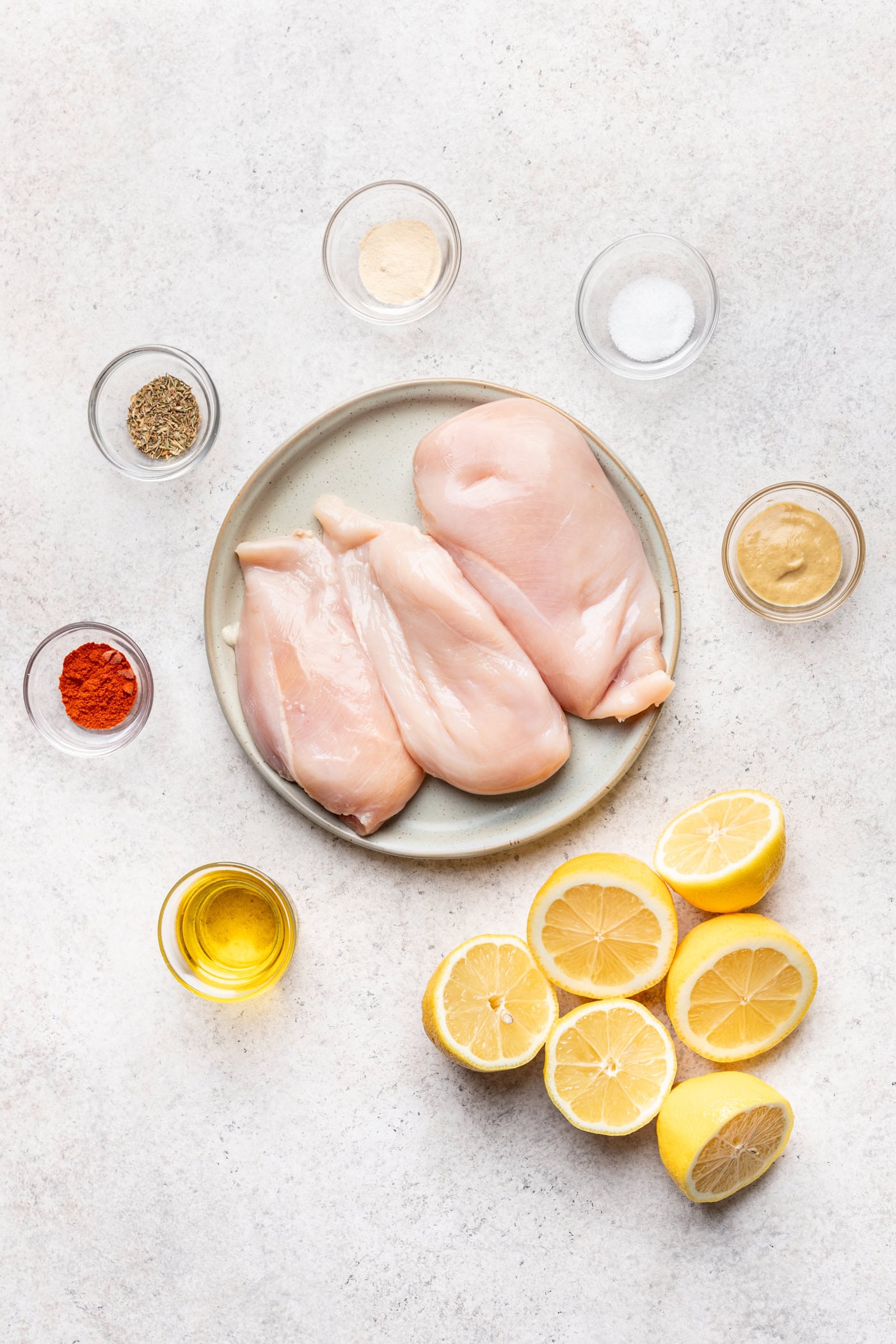 A plate with two raw chicken breasts, surrounded by halved lemons, small bowls of mustard, olive oil, paprika, black pepper, garlic powder, and salt on a light surface.