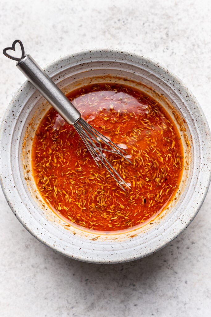 A ceramic bowl filled with an orange-red seasoned liquid marinade, with visible herbs and spices, and a metal whisk with a heart-shaped handle resting inside. The bowl is on a light, textured surface.