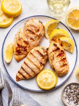 Three grilled chicken breasts on a plate, garnished with lemon slices. Surrounding the plate are more lemon slices, a small bowl of salt and pepper, a glass bottle of oil, and silver forks on a napkin.