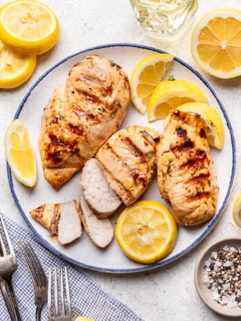 A plate with grilled chicken breasts, some sliced and some whole, garnished with lemon slices. Surrounding the plate are lemon halves, forks, a napkin, and a small bowl of salt and pepper.