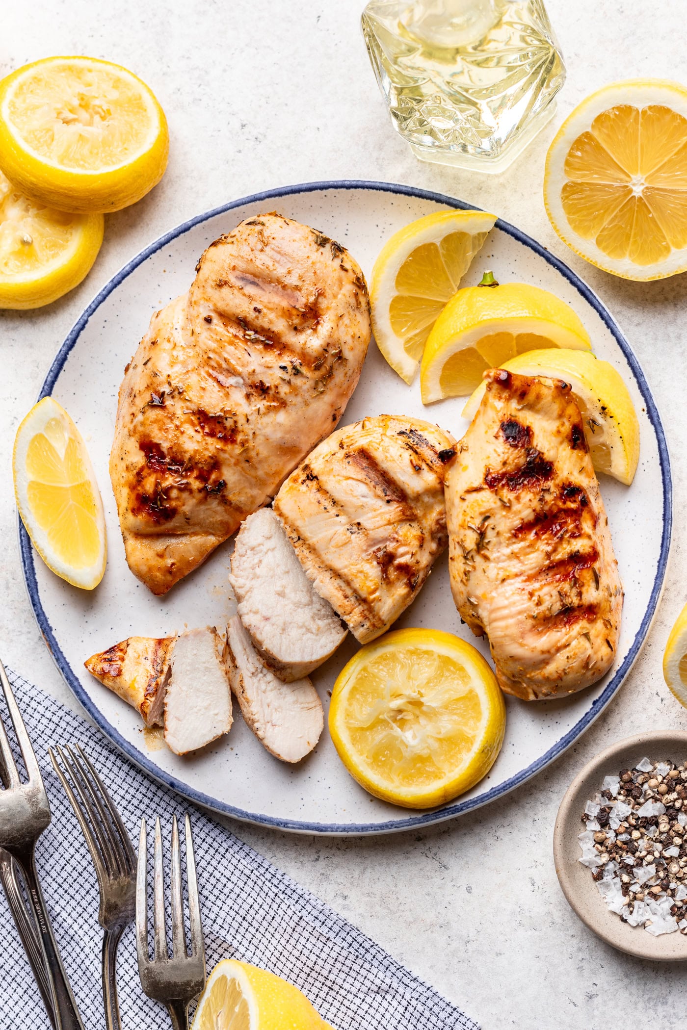 A plate with grilled chicken breasts, some sliced and some whole, garnished with lemon slices. Surrounding the plate are lemon halves, forks, a napkin, and a small bowl of salt and pepper.