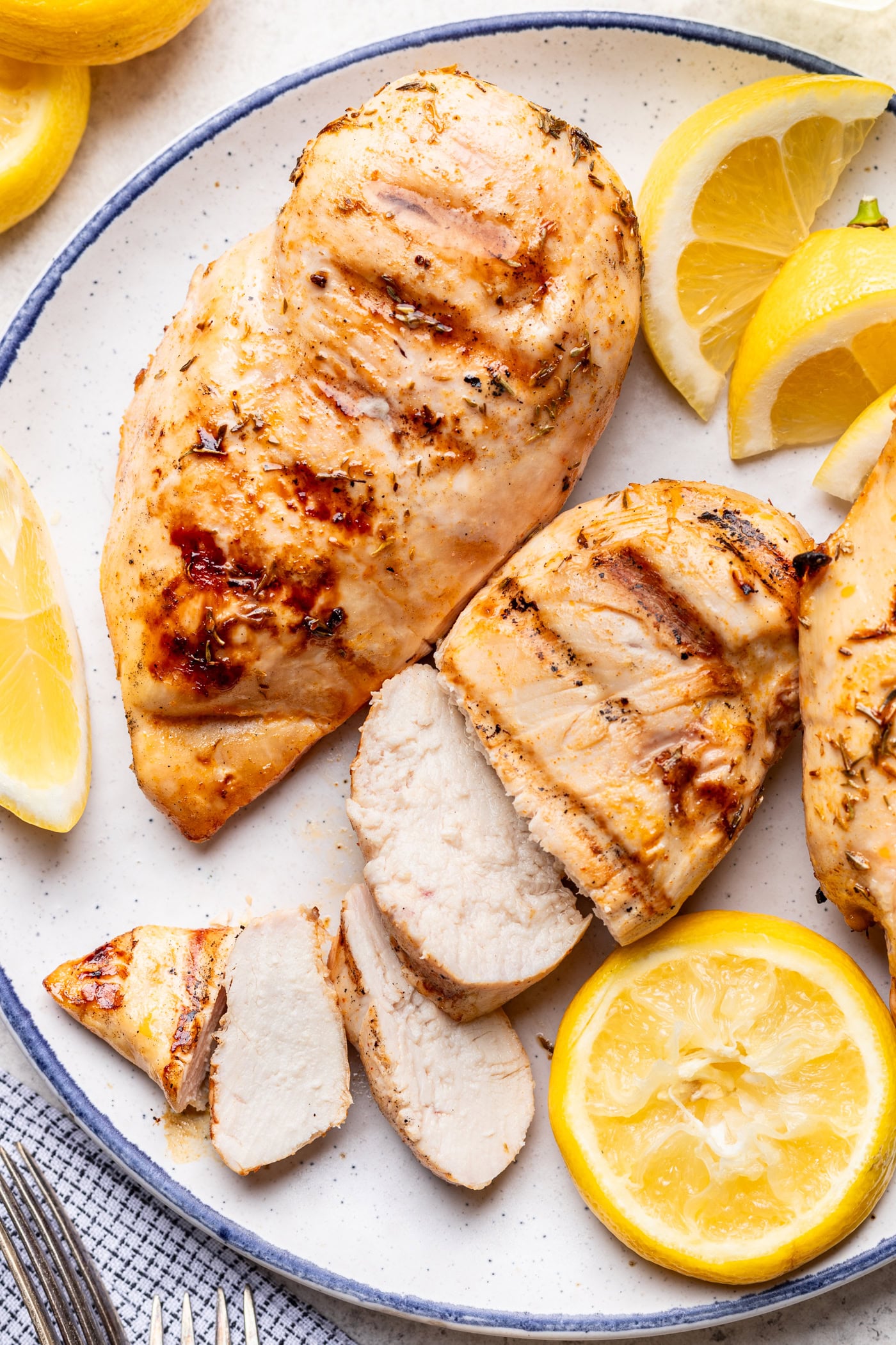 Grilled chicken breast on a white plate, some pieces sliced, served with lemon wedges and a slice of lemon. The chicken has grill marks and a golden brown color.