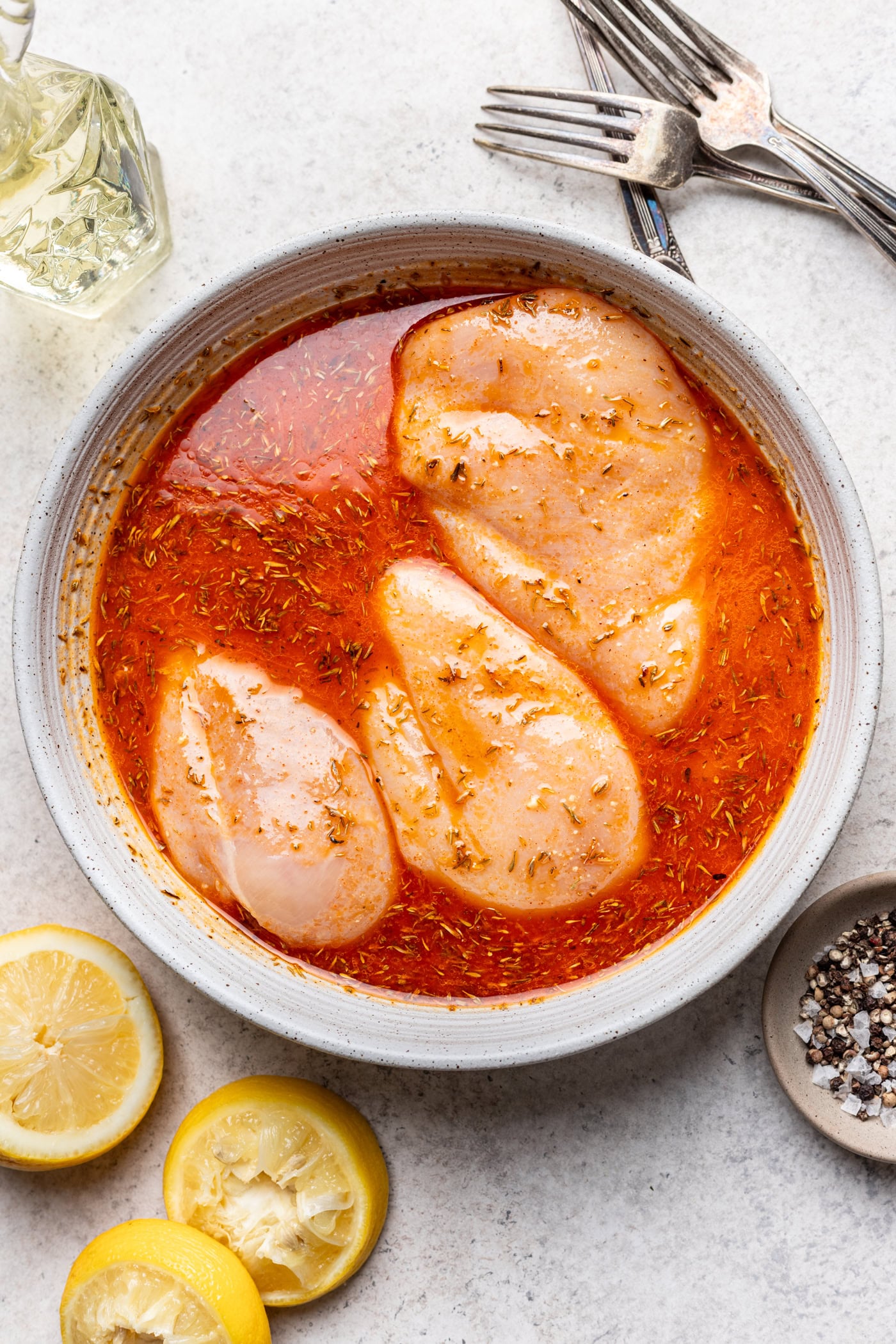 Raw chicken breasts marinating in a red, seasoned liquid in a bowl, surrounded by lemon halves, a bowl of pepper, two forks, and a glass container of oil on a light countertop.