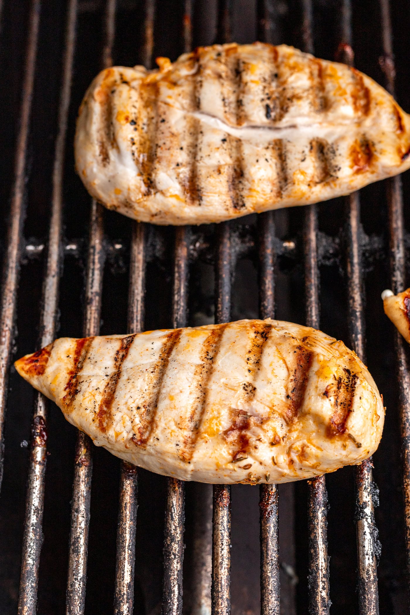 Two pieces of grilled chicken breast with visible grill marks are cooking on a barbecue grill. The chicken is lightly browned and the grill grates are blackened from use.