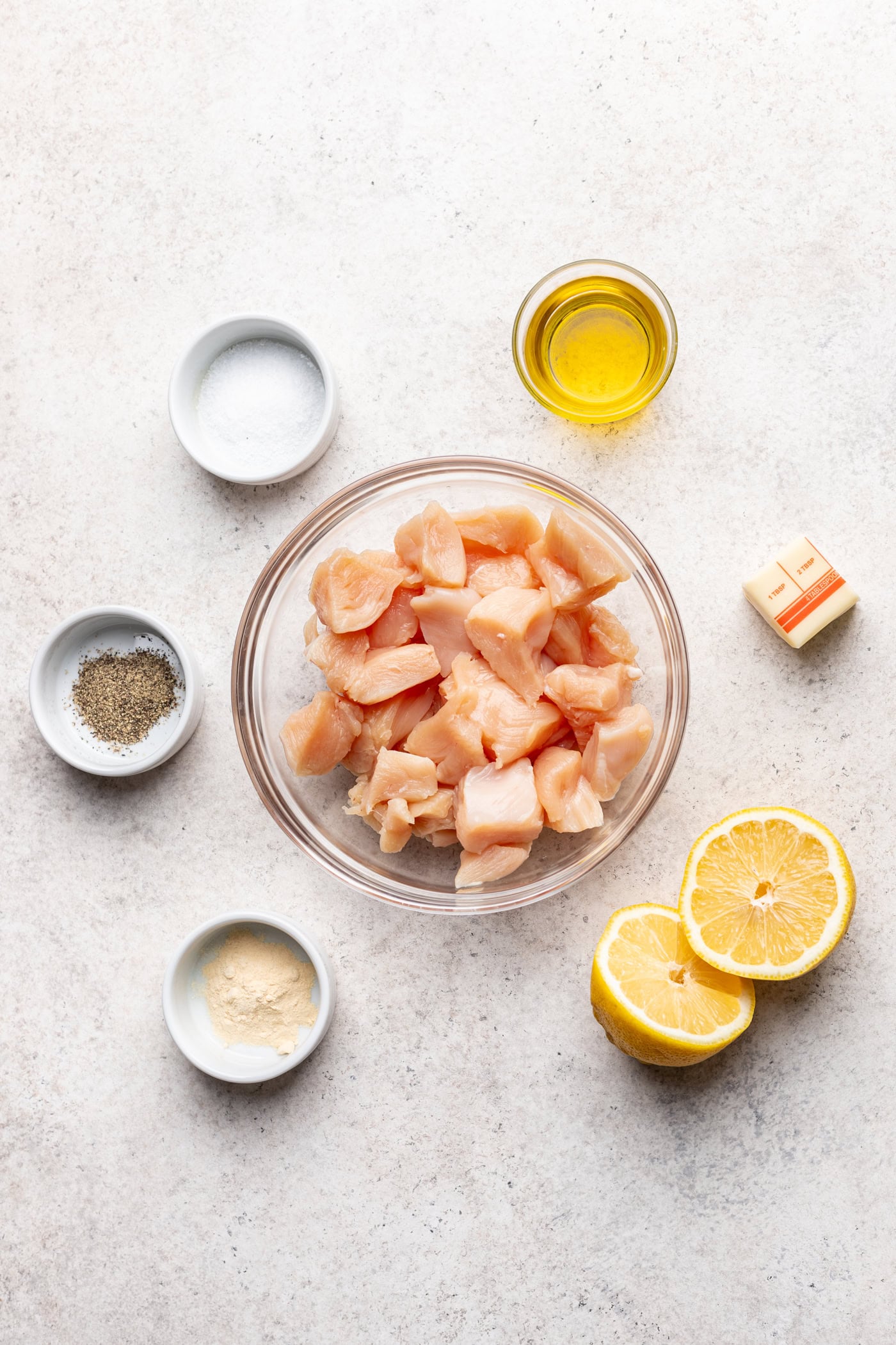 Raw diced chicken breast in a glass bowl, surrounded by salt, pepper, garlic powder, olive oil, butter, and a halved lemon on a light countertop.