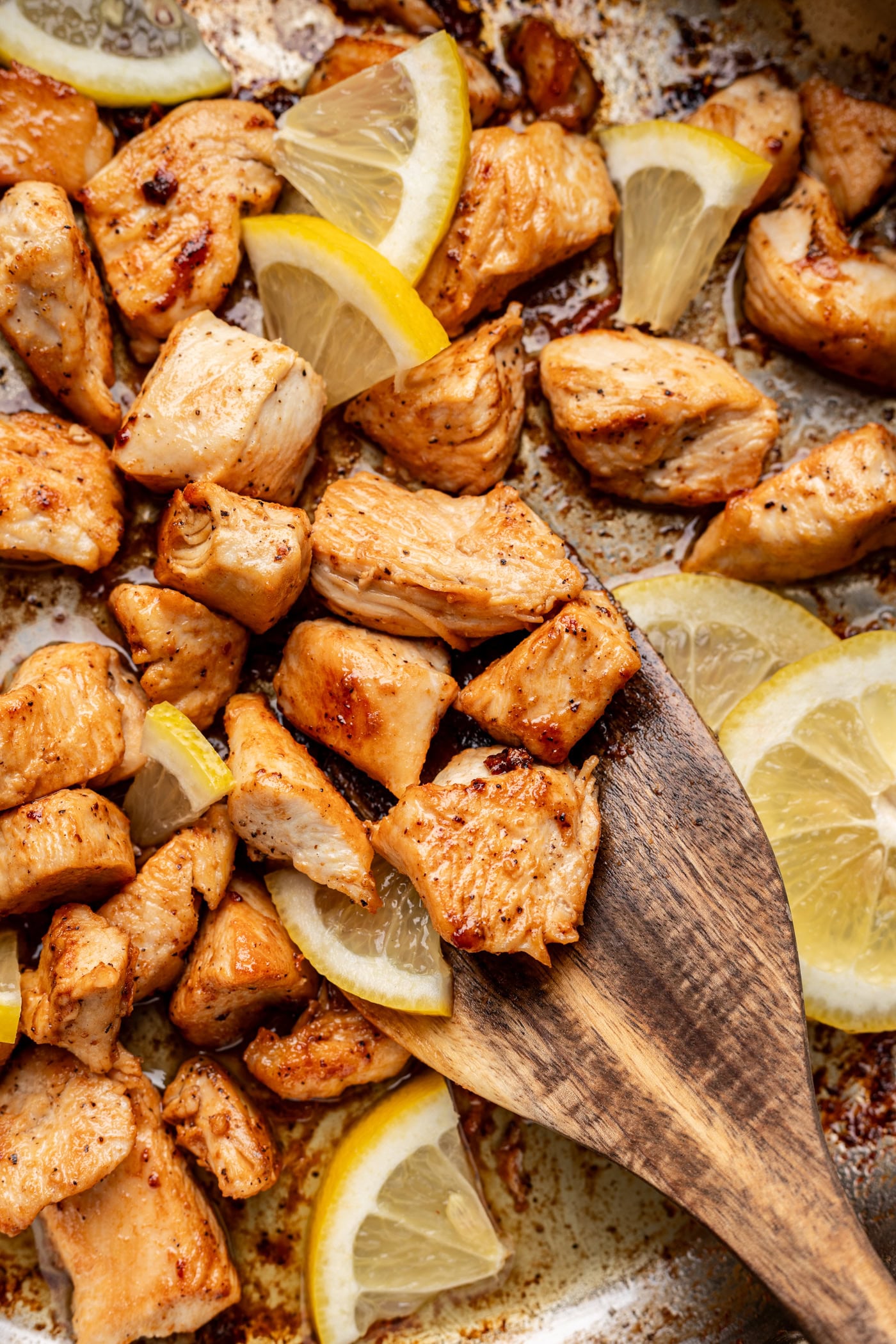 Cooked, browned chunks of chicken are mixed with lemon wedges in a pan. A wooden spatula lifts some pieces, showing the juicy texture and seasoning on the chicken.