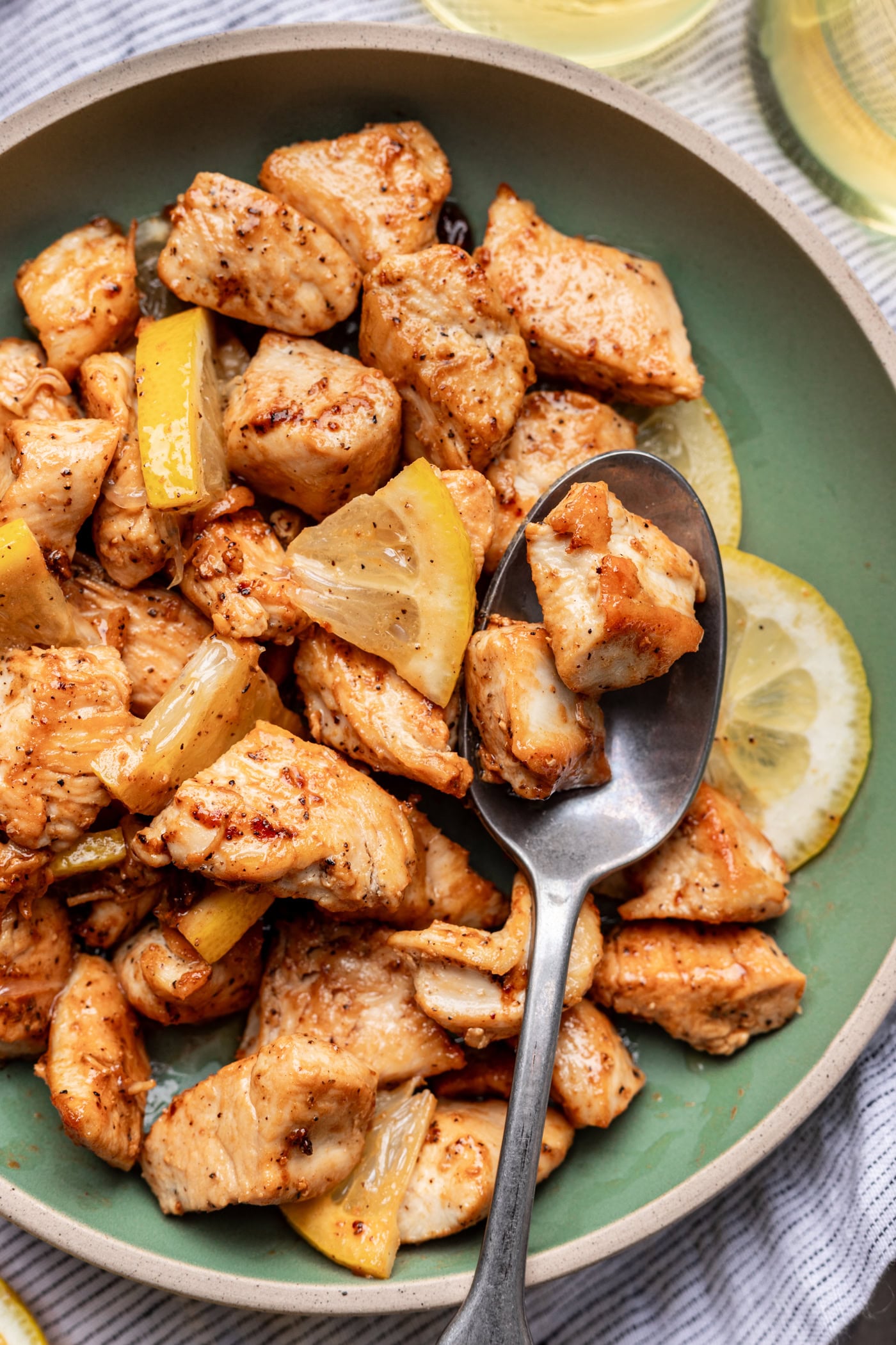 A bowl of cooked chicken pieces mixed with lemon slices. A spoon is scooping up some chicken, and the dish sits on a striped cloth. The chicken looks seasoned and juicy.