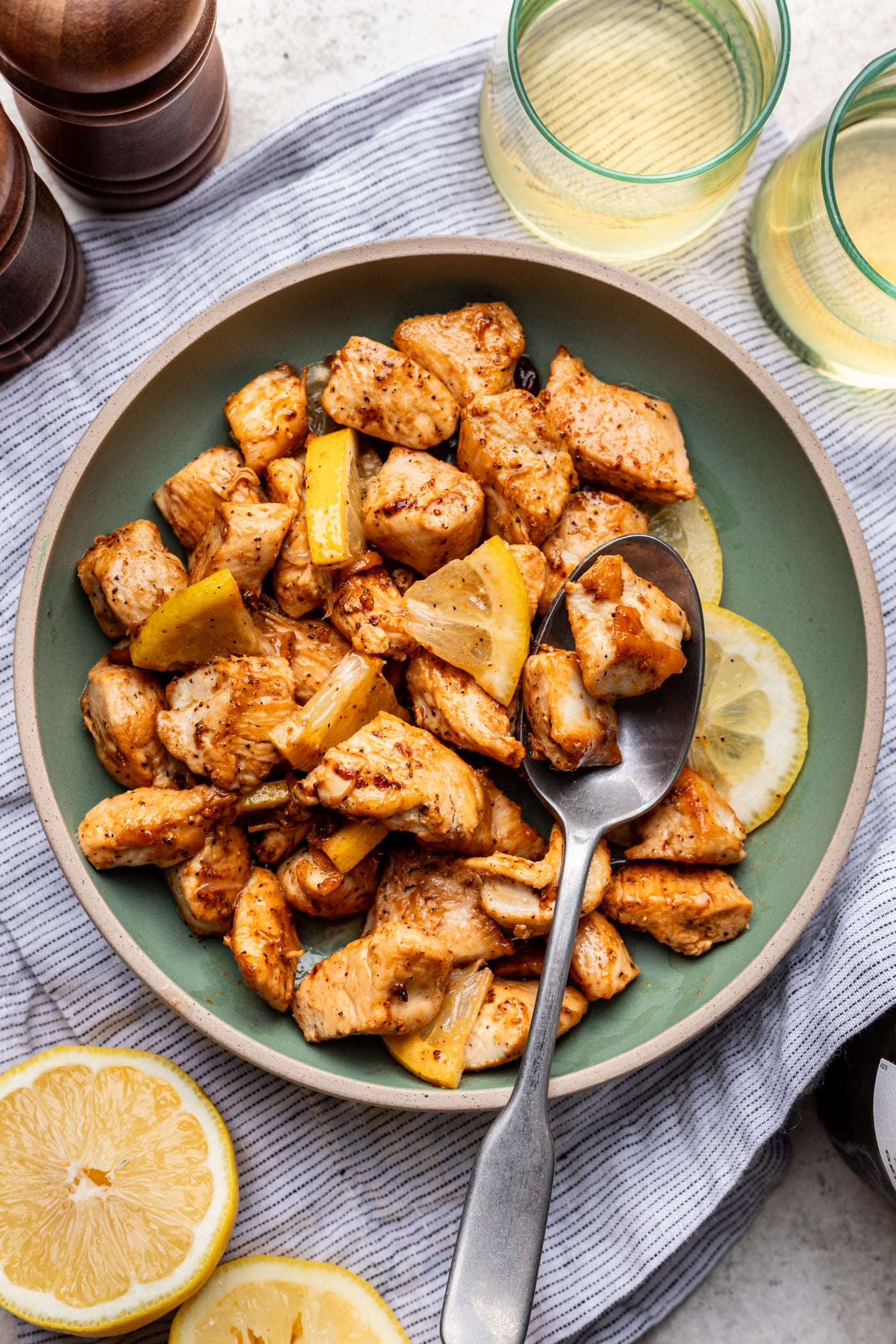 A bowl of cooked, seasoned chicken pieces with lemon wedges, a spoon, and slices of lemon on the side, next to glasses of a light beverage and a striped cloth.
