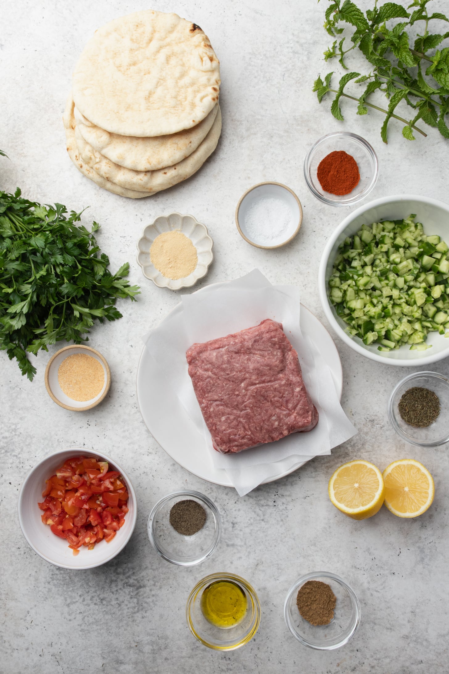 pita, raw ground meat, diced cucumber, chopped tomato, fresh herbs, lemon halves, olive oil, and assorted spices in small bowls arranged on a white surface.