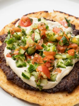 A flatbread topped with a layer of creamy white sauce, minced grilled meat, and a fresh tomato-cucumber salad, served on a white plate.