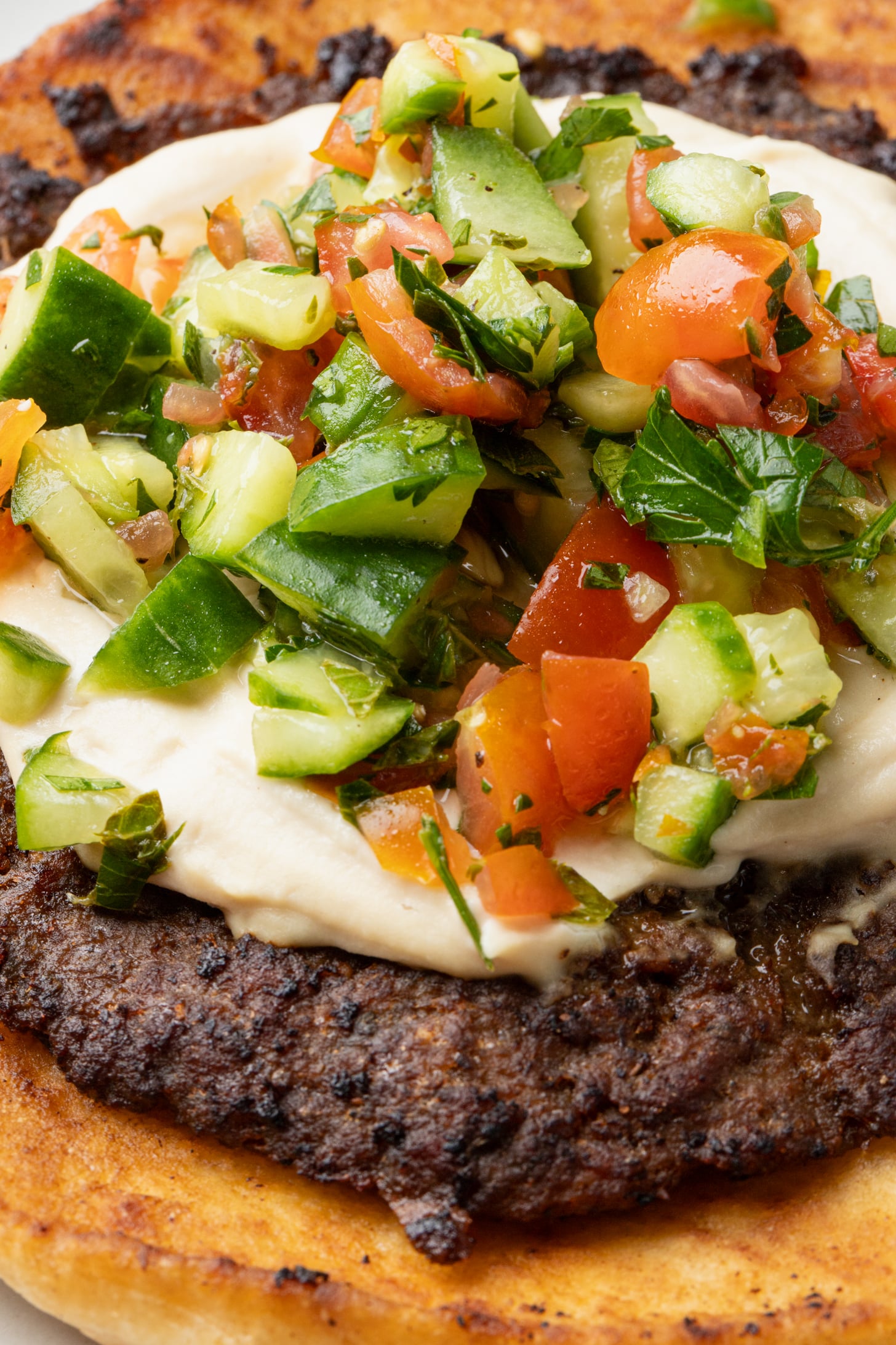 Close-up of a flatbread topped with grilled ground meat, a creamy white sauce, and a colorful chopped salad of cucumbers, tomatoes, and herbs.