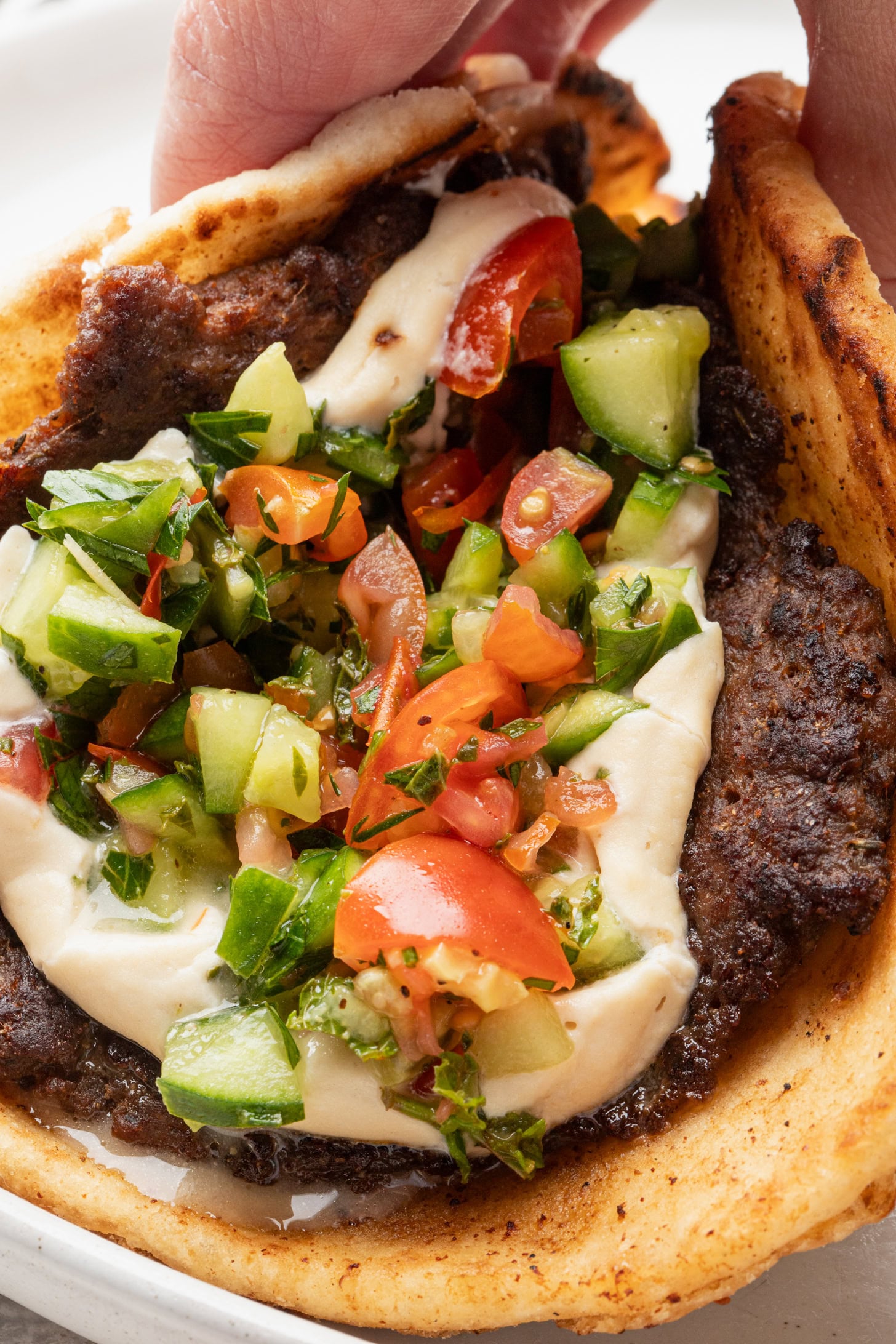 A close-up of a hand holding a pita bread filled with grilled meat, creamy sauce, and a fresh tomato and cucumber salad.