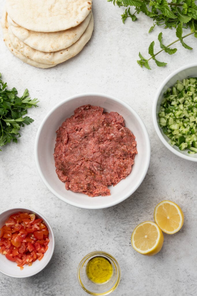 A bowl of raw ground meat surrounded by pita bread, fresh herbs, chopped cucumber, diced tomato, lemon halves, and a small dish of olive oil on a light countertop.