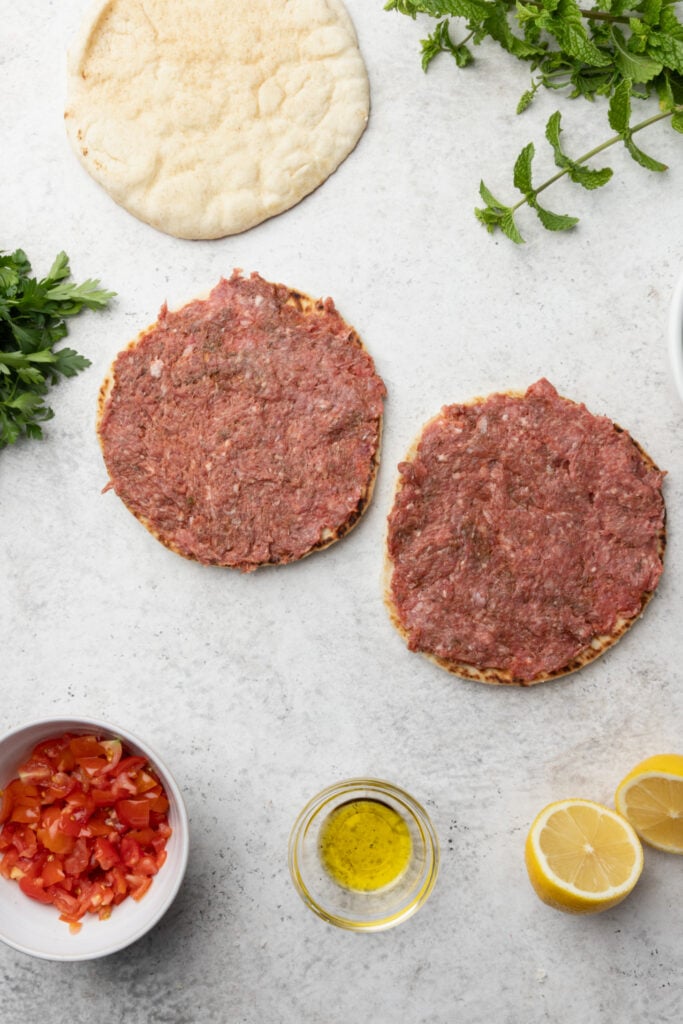 Two flatbreads topped with raw minced meat are on a white surface, surrounded by chopped herbs, a pita, diced tomatoes in a bowl, olive oil, and halved lemons.
