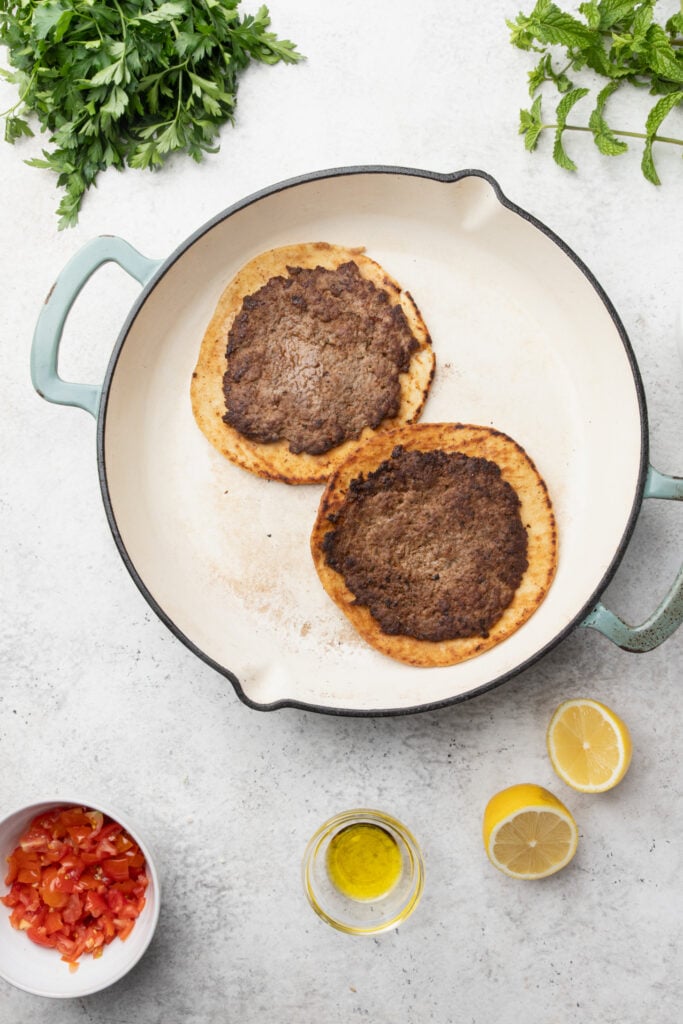 A white pan with two pieces of flatbread topped with ground meat. Surrounding the pan are fresh herbs, a bowl of chopped tomatoes, a halved lemon, and a small dish of olive oil.