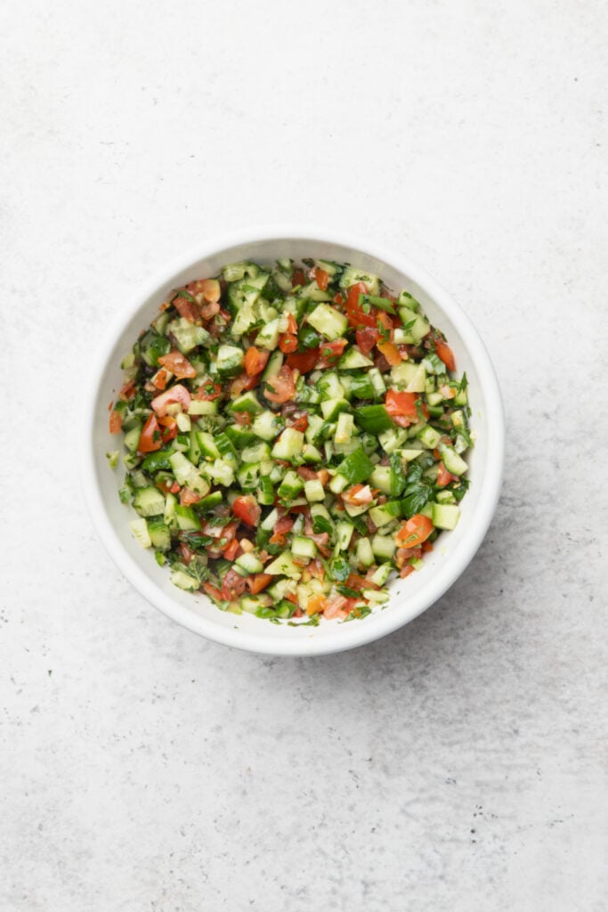 A white bowl filled with freshly chopped salad, including diced tomatoes, cucumbers, herbs, and green peppers, on a light gray surface.