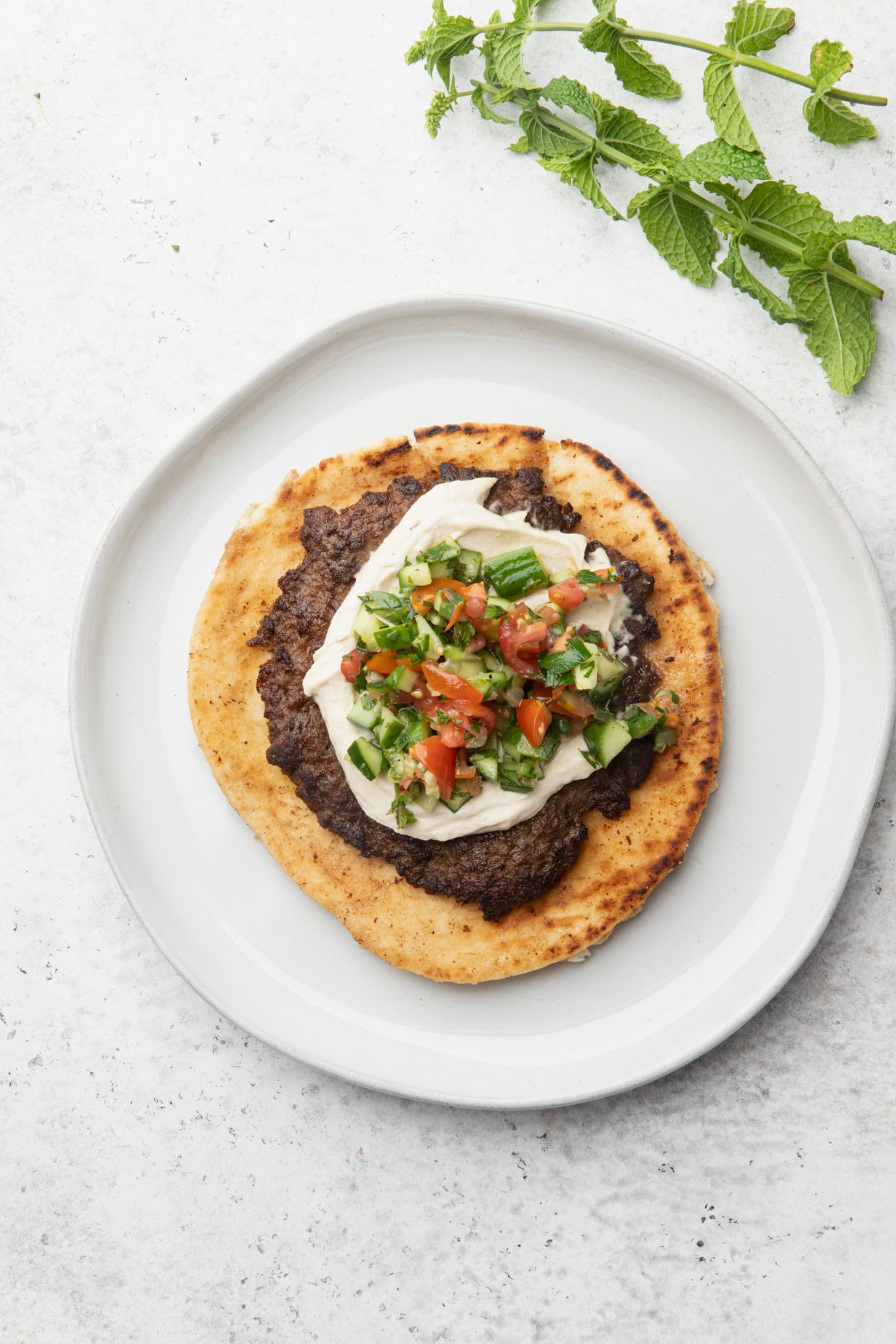 Flatbread topped with ground meat, creamy white sauce, and chopped tomato, cucumber, and herbs sits on a white plate. Fresh mint and parsley sprigs are placed above the plate on a light background.