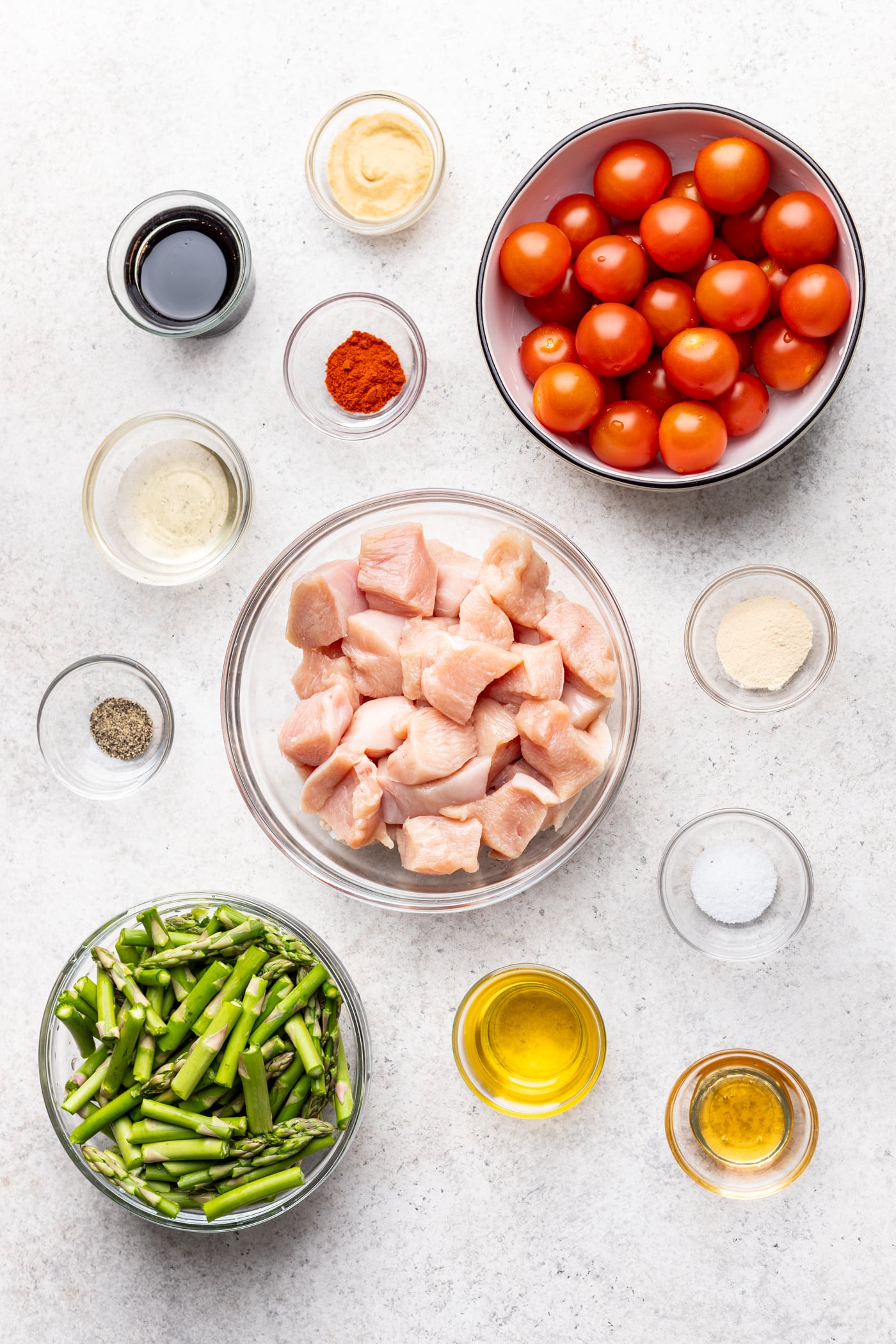 Top-down view of bowls with raw chicken cubes, cherry tomatoes, chopped asparagus, olive oil, seasonings, soy sauce, mustard, and seasoning powders arranged on a light surface.