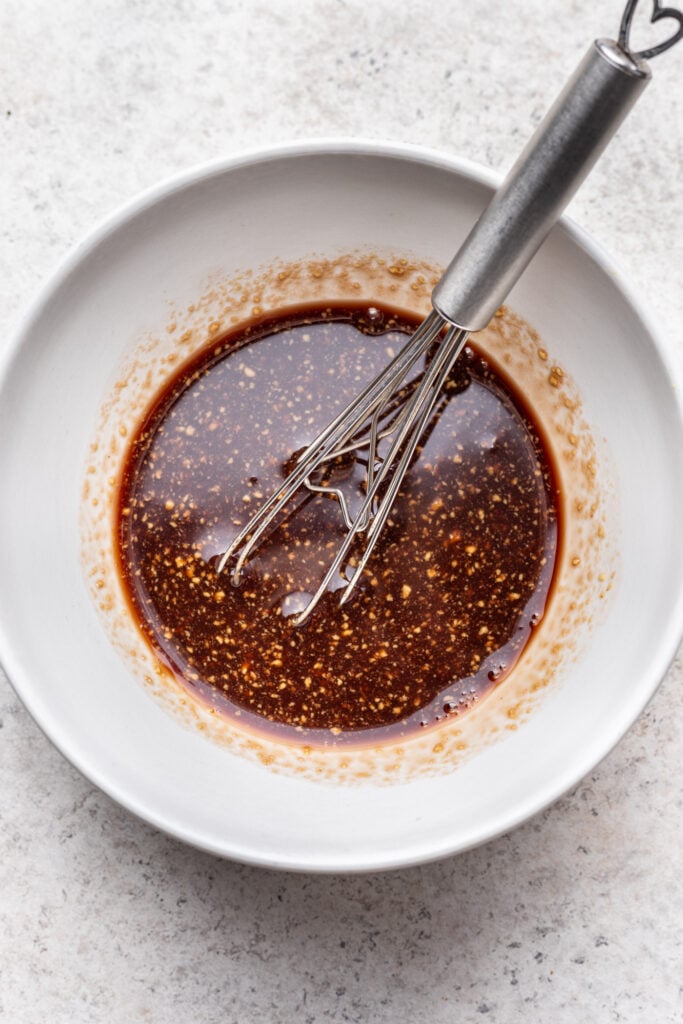 A metal whisk rests in a white bowl filled with a dark, speckled sauce, likely a marinade or dressing, on a light textured surface.