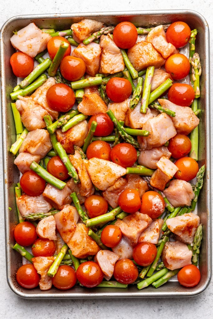 A baking tray filled with raw diced chicken, cherry tomatoes, and chopped asparagus, all seasoned and ready to be cooked.