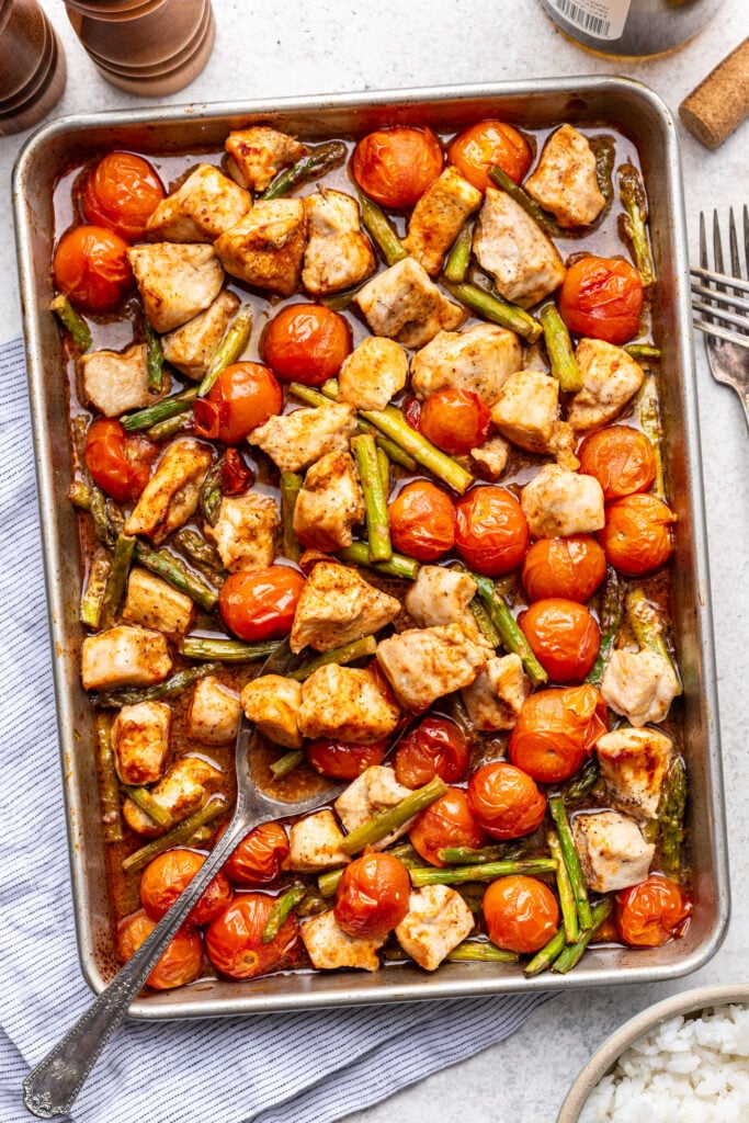 Baked chicken pieces, cherry tomatoes, and green beans on a sheet pan with a serving spoon, placed on a table next to a blue striped towel, two forks, and a bowl of white rice.