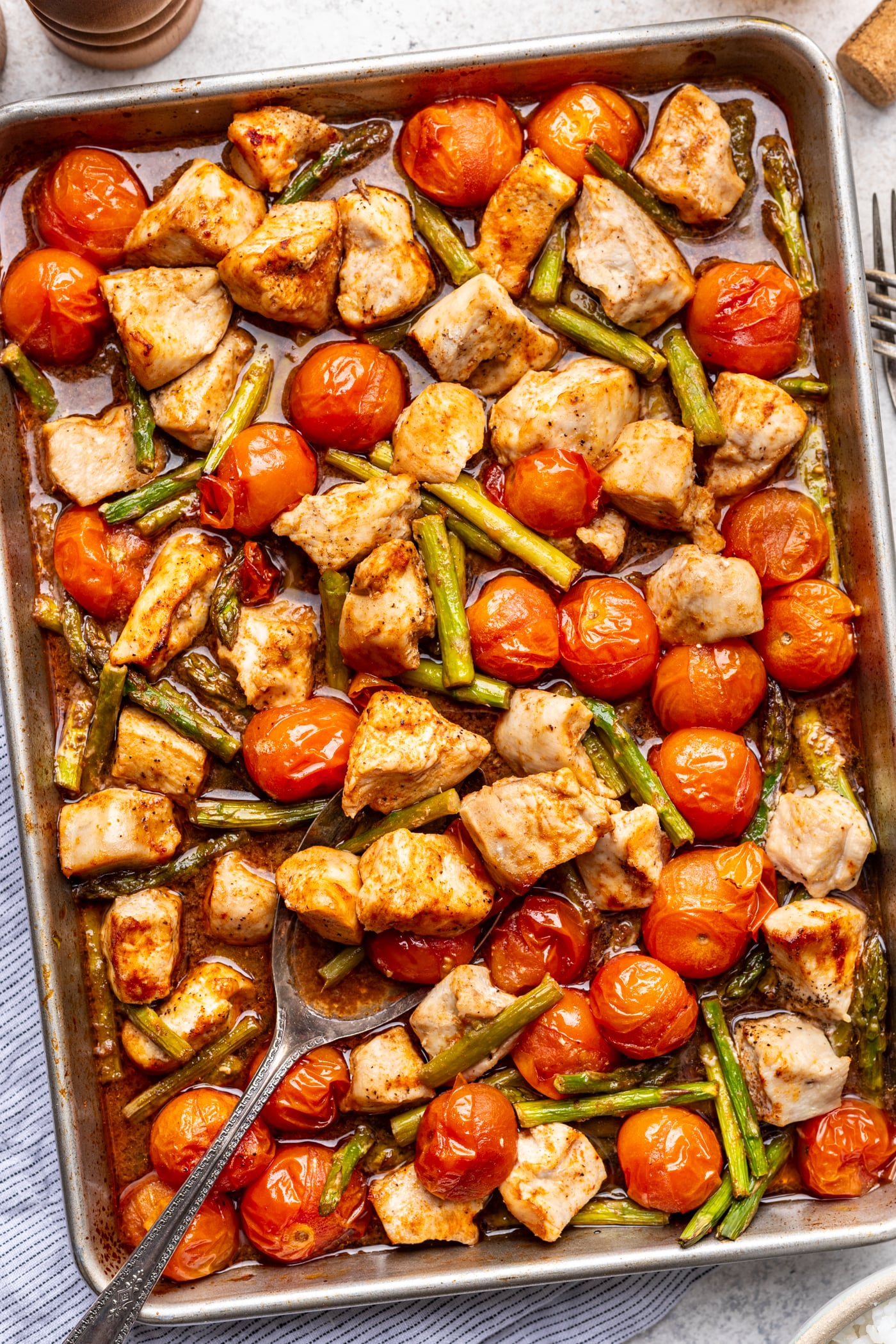 A sheet pan filled with cooked chunks of chicken, bright red cherry tomatoes, and green beans, all seasoned and roasted, with a serving spoon resting on the pan.