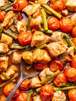 A spoon scoops up chunks of cooked chicken, cherry tomatoes, and green beans, all roasted together and coated in seasoning.
