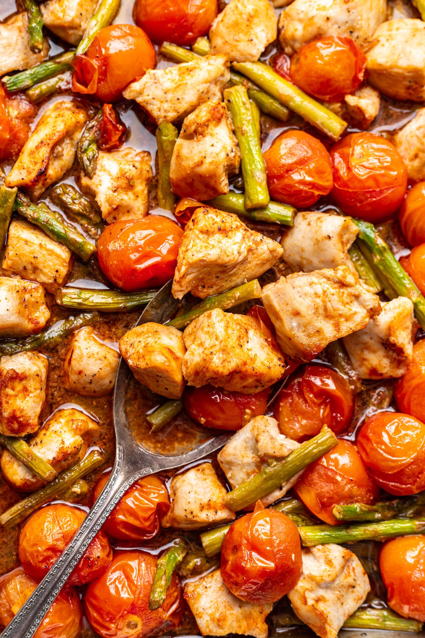 A spoon scoops up chunks of cooked chicken, cherry tomatoes, and green beans, all roasted together and coated in seasoning.