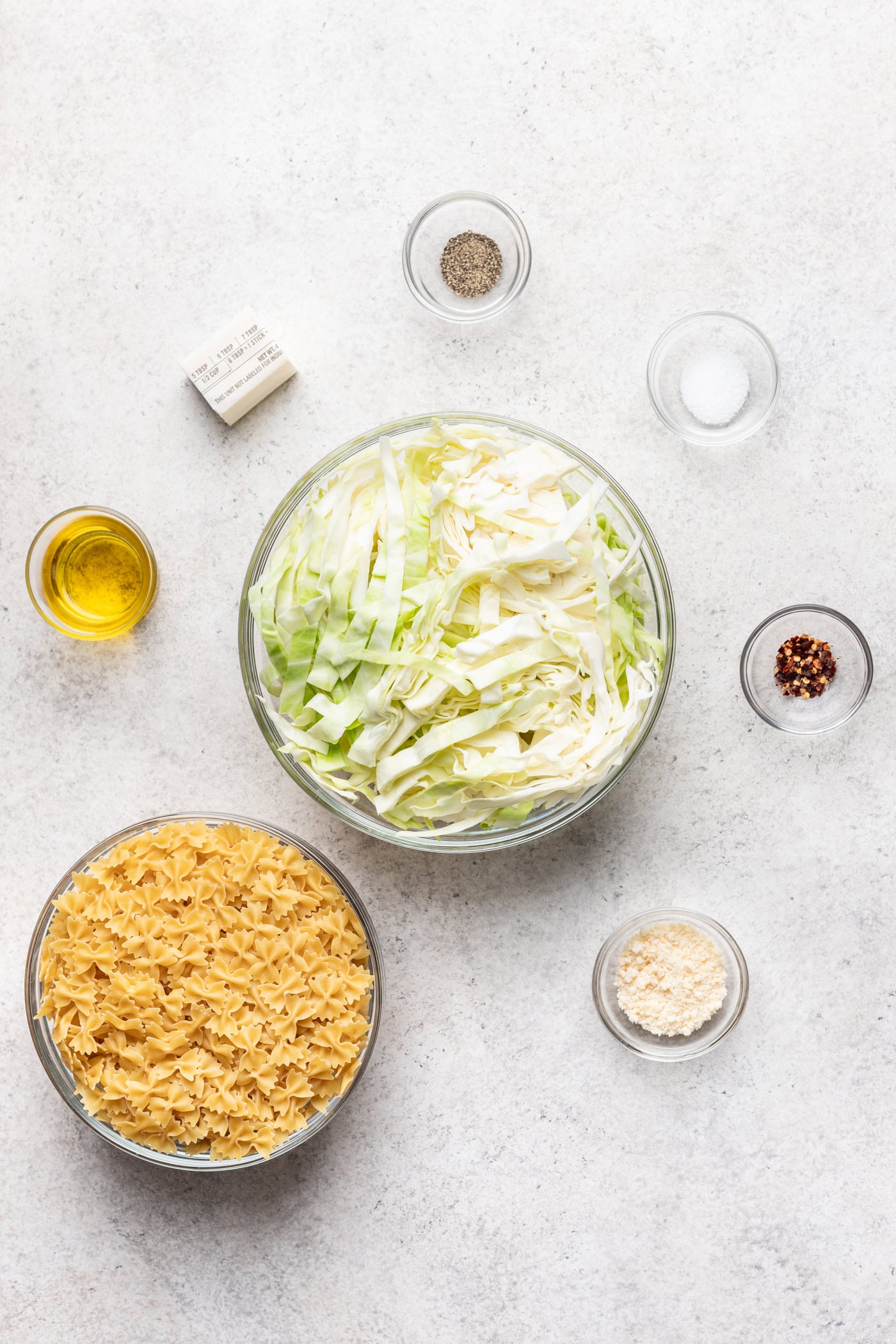 Bowls of uncooked egg noodles, shredded cabbage, grated cheese, oil, butter, salt, pepper, and red pepper flakes are arranged on a light surface, ready for preparation.