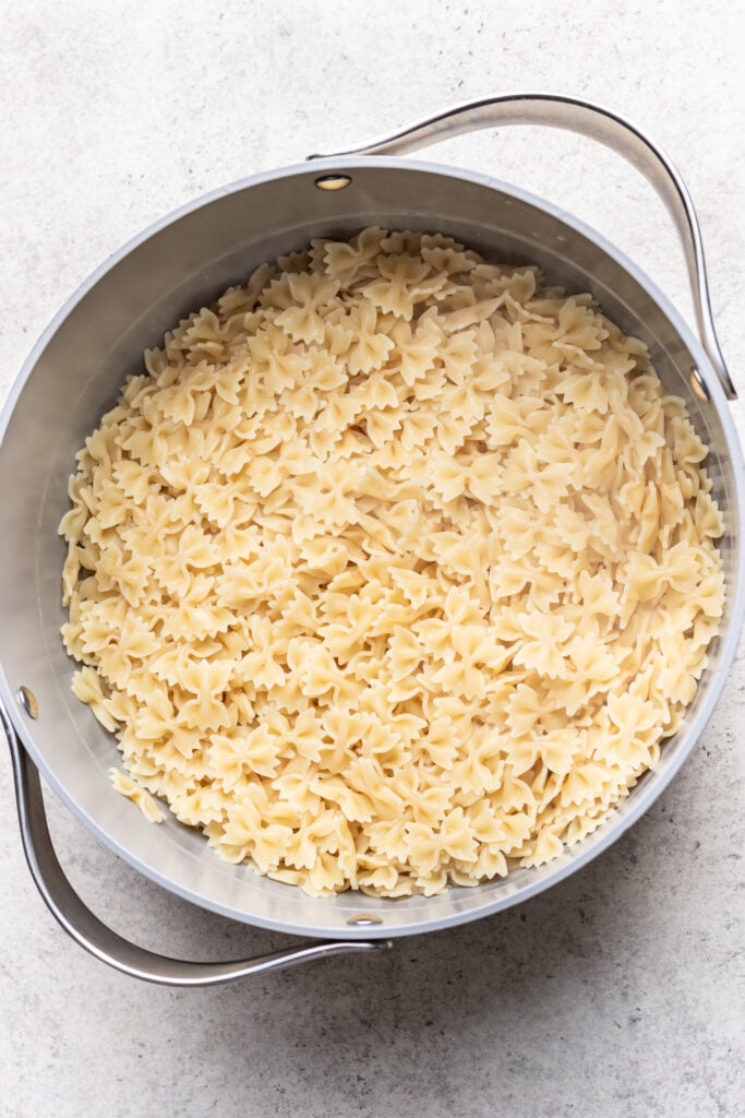 A large metal pot filled with cooked farfalle pasta, also known as bow-tie pasta, sits on a light-colored surface.