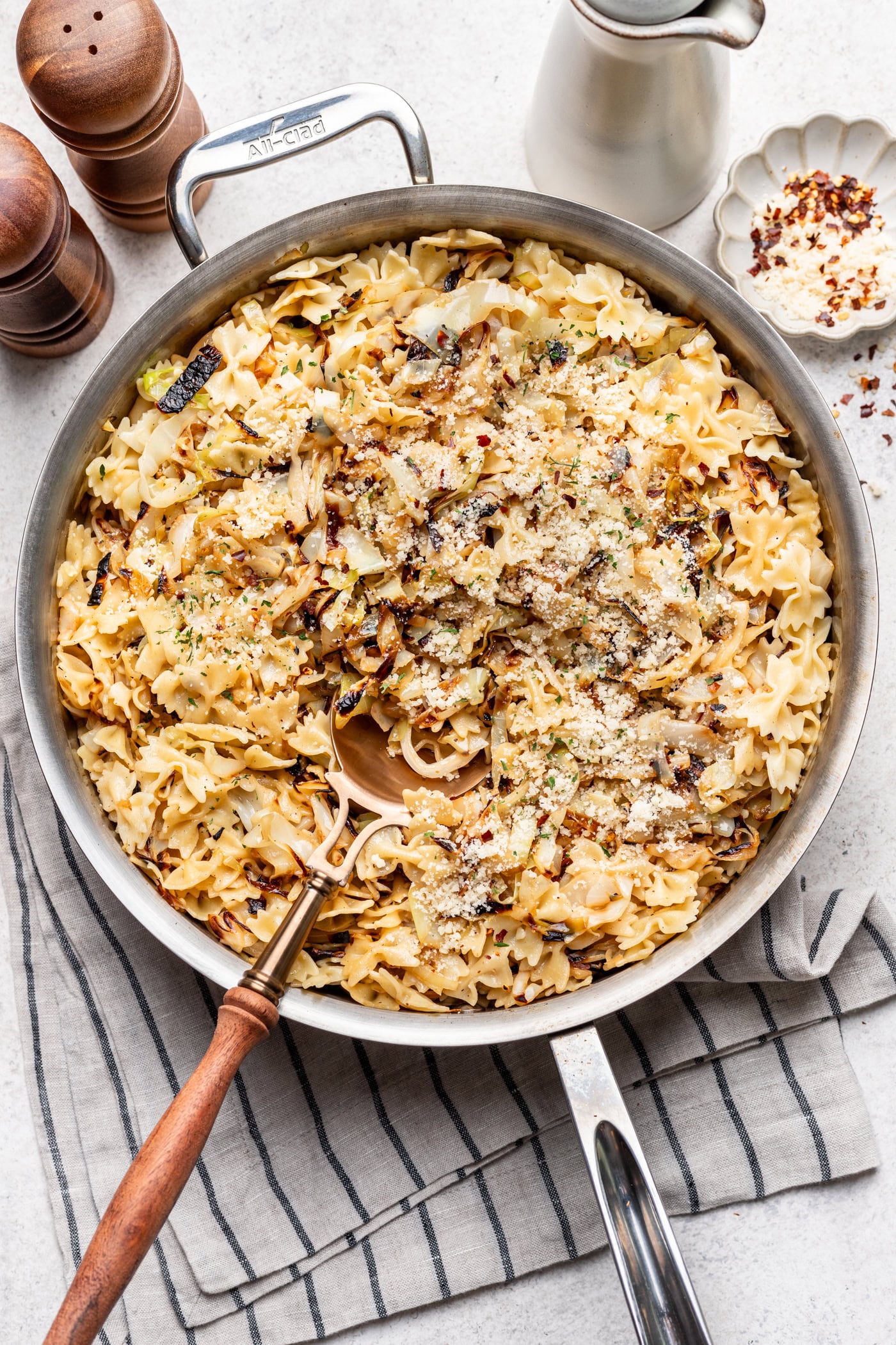 A skillet filled with bowtie pasta mixed with caramelized cabbage, topped with grated cheese and herbs, sits on a striped cloth, with a wooden spoon, salt and pepper shakers, and a small dish of red pepper flakes nearby.