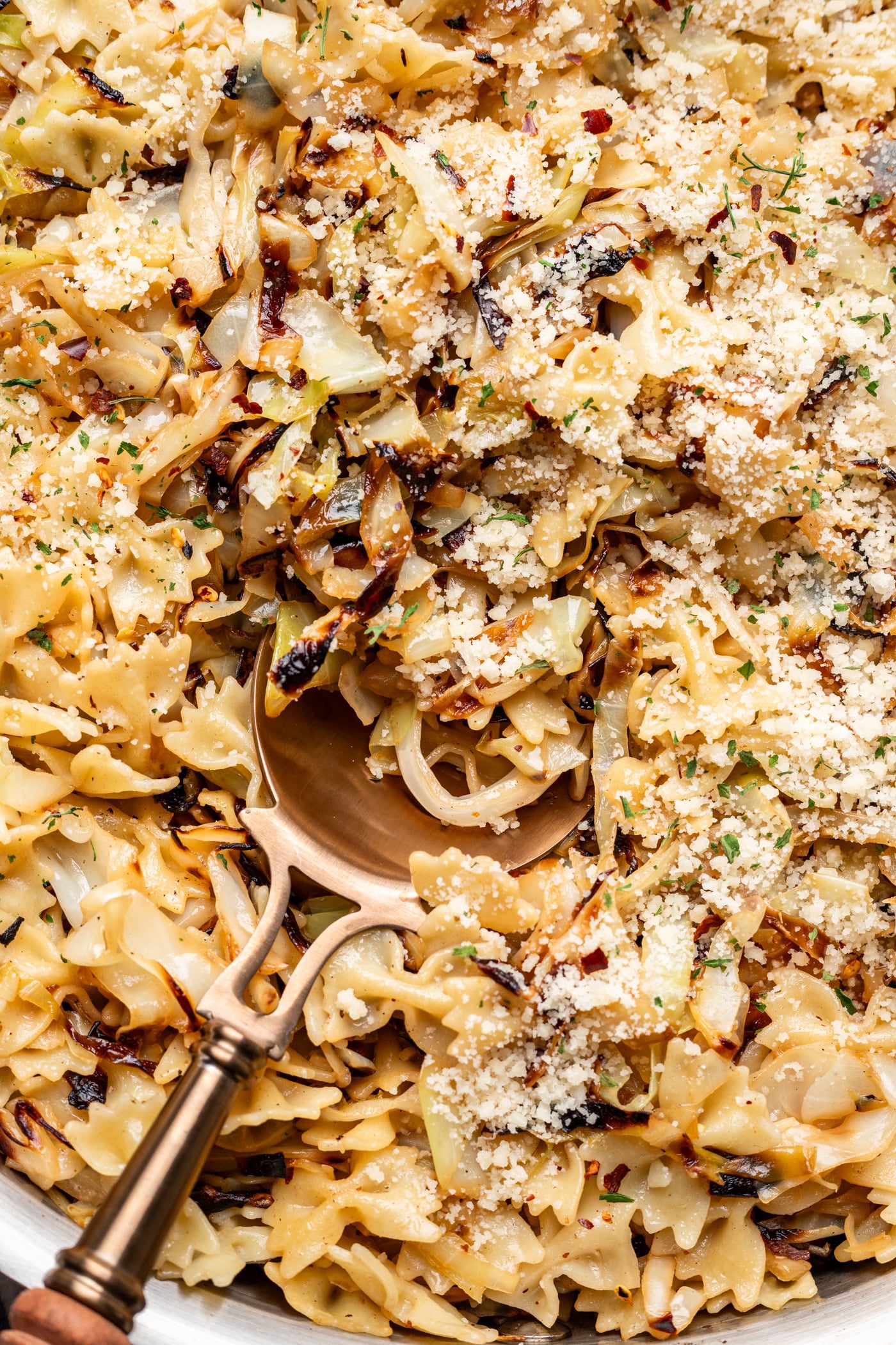 Close-up of bow tie pasta mixed with caramelized cabbage, sprinkled with grated cheese and herbs. A large serving spoon rests in the pasta, ready to serve.