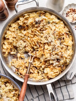 A skillet of bowtie pasta with caramelized cabbage sits on a striped towel. A wooden-handled fork rests in the pasta. Pepper mills, a jug, and a dish of red pepper flakes are nearby.