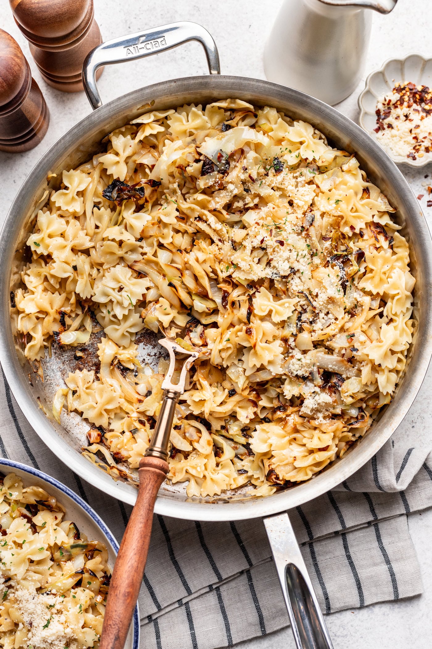 A skillet of bowtie pasta with caramelized cabbage sits on a striped towel. A wooden-handled fork rests in the pasta. Pepper mills, a jug, and a dish of red pepper flakes are nearby.