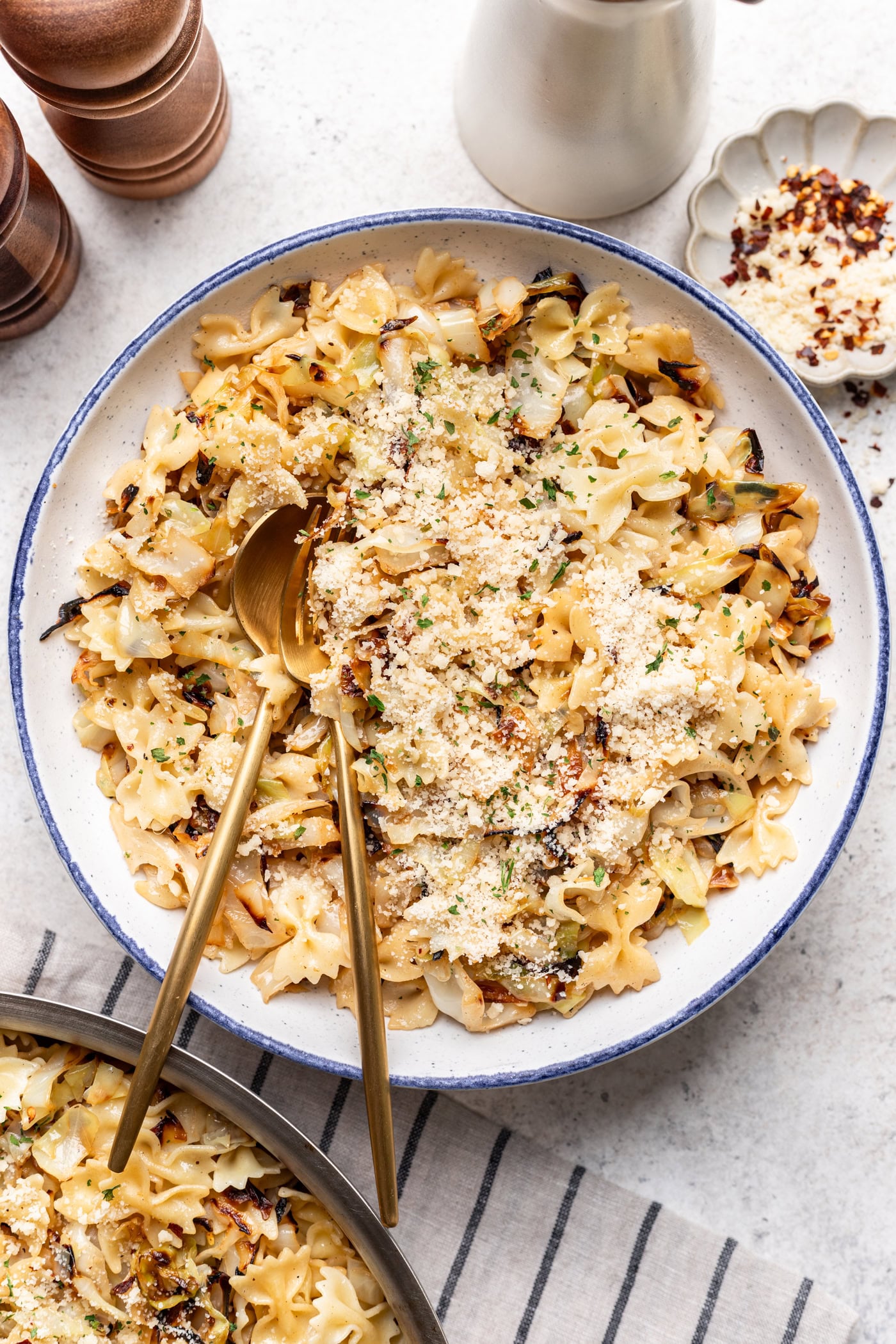 A bowl of bow tie pasta mixed with roasted cauliflower, topped with grated cheese and herbs, with two gold utensils. Pepper mills, a sauce dish, and a napkin are nearby.
